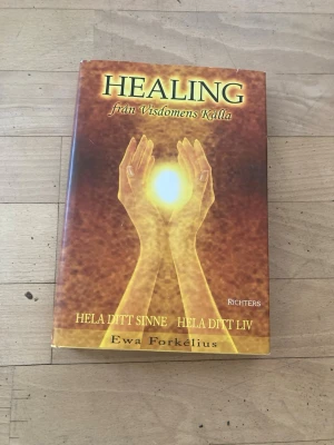 Healing från visdomens källa - En spirituell bok av Ewa Forkelius med titeln 'Healing från visdomens källa'. Omslaget är brunt och gult med motiv av två händer som håller ett lysande ljus. Perfekt för dig som är intresserad av healing, personlig utveckling och inre balans.