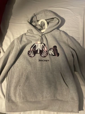 Grå Palm Angels hoodie - Snygg grå hoodie från Palm Angels med ikoniskt björntryck och logga på bröstet. Stor Palm Angels-text i svart på ryggen. Klassisk känguruficka och huva med dragsko. Perfekt för dig som gillar streetwear och vill sticka ut. Aldrig använd.