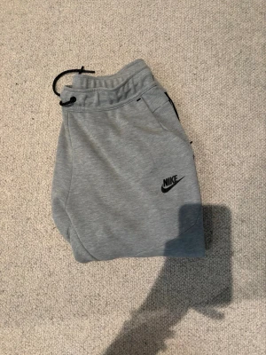 Grå mjukisbyxor från Nike - Gråa mjukisbyxor från Nike med svart logga på vänster ben. Byxorna har dragsko i midjan och sidofickor. Tillverkade i mjukt och bekvämt material, perfekta för chill eller träning. Snygg och enkel design som funkar till det mesta.
