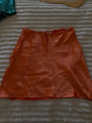 Orange satin kortkjol - Säljer en snygg orange kortkjol i glansigt satinmaterial. Kjolen har en enkel, rak passform.