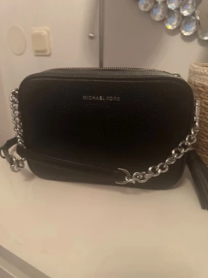 Svart axelväska Michael Kors aldrig använd - Snygg svart axelväska från Michael Kors i rektangulär modell. Väskan har silverfärgad kedja och dragkedja upptill samt logga framtill. Tillverkad i läder med stilren design, perfekt för att bära det viktigaste.