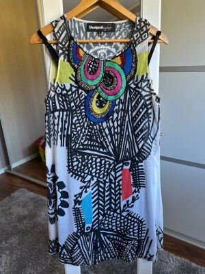 Mönstrad klänning från Desigual - Färgstark och unik klänning från Desigual med svartvitt grafiskt mönster och broderade detaljer i grönt, rosa, blått och gult vid halsen. Ärmlös modell i lätt material, perfekt för dig som gillar att sticka ut med din stil.