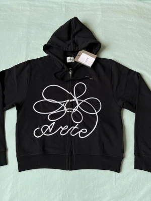 Svart hoodie med vit logga från Arte - Svart hoodie från Arte med dragkedja och stor vit grafisk logga på framsidan. Klassisk huva med snörning och ribbade muddar. Perfekt för en streetwear-look och enkel att matcha med jeans eller joggers.