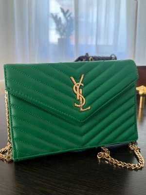 Grön kuvertväska från YSL i läder - Snygg grön kuvertväska från YSL i quiltat läder med guldigt YSL-emblem framtill. Väskan har en elegant kedjerem i guld och klassisk kuvertform. Perfekt för dig som vill sticka ut med en färgstark accessoar.