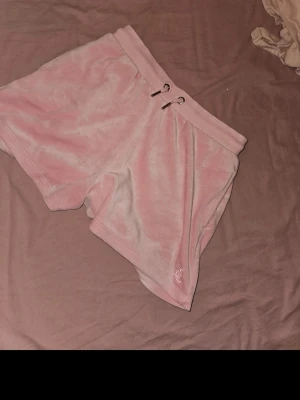 Rosa velourshorts med snörning - Mysiga rosa shorts i velour med bred resår i midjan och snörning framtill. Shortsen har en bakficka och en liten broderad detalj på ena benet. Perfekta för chill dagar eller när du vill vara extra comfy. Storleken motsvarar en XS/S