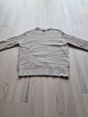 Beige stickad tröja med strukturmönster - Säljer en beige stickad tröja med snyggt strukturmönster och rund hals. Tröjan har långa ärmar och ribbade muddar vid ärmslut och nederkant. Perfekt för dig som gillar en stilren och enkel look.