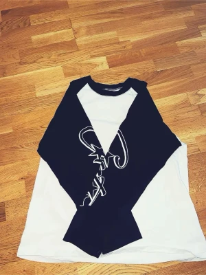 Svartvit långärmad topp med tryck Corteiz oversize - Cool långärmad topp från Corteiz i svart och vitt med kontrastärmar och grafiskt tryck på ärmarna. Klassisk baseballmodell med rund halsringning och relaxed fit. Perfekt för en avslappnad streetstyle-look.