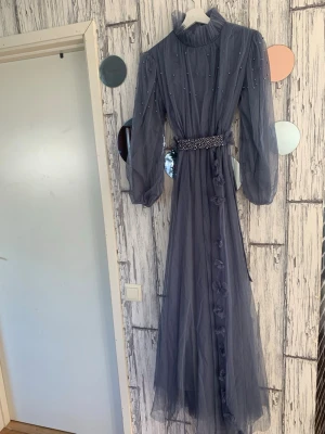 Lång blå tyllklänning med pärlor - Elegant långklänning i blå tyll med pärldetaljer på överdelen och ärmarna. Klänningen har hög krage, långa puffärmar och ett markerat midjebälte dekorerat med pärlor. Framtill finns dekorativa blommor längs kjolen för en extra romantisk touch.