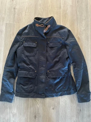 Mörkblå fieldjacka från Massimo dutti - Snygg mörkblå filed jacka med fyra framfickor och hög krage med spänne. Jackan har justerbara band i midjan och tryckknappar på fickorna. Perfekt för dig som gillar en stilren och funktionell look.