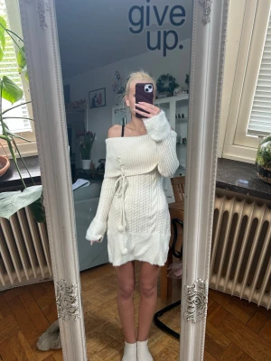 Vit stickad offshoulder klänning - Supermysig vit stickad klänning med offshoulder och kabelstickat mönster. Klänningen har snörning i midjan med söta tofsar och långa ärmar. Perfekt för dig som vill ha en söt och trendig look!