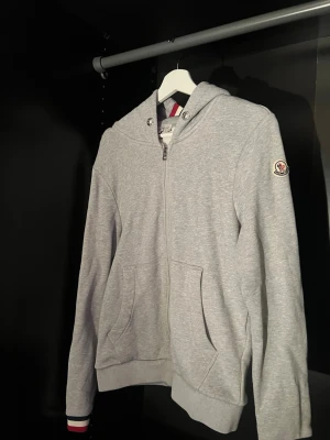 Grå Moncler hoodie med dragkedja - Fungerande nfc tag, tröjan har lite defekter som några små hål i slutet av ärmen och ett på ena armbågen. Hoodien är storlek S men sitter lite mellan S och XS. Kom med frågor och förslag på pris, kan skicka mer detaljerade bilder om ni frågar✅