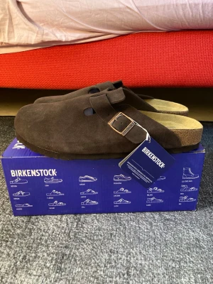Birkenstock Boston tofflor mocha - Säljer ett par Birkenstock Boston tofflor i färgen mocha. De är tillverkade i mocka med en justerbar rem och metallspänne. Sulan är ergonomiskt utformad för extra komfort. Perfekta för dig som gillar stilrena och bekväma skor.