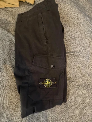 Svarta cargobyxor från Stone Island - Snygga svarta cargobyxor från Stone Island med klassisk logotyp-patch på benfickan. Byxorna har flera fickor, raka ben och är tillverkade i ett slitstarkt bomullsmaterial. Perfekta för en streetwear-look.