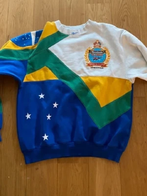 Reebok sweatshirt med brodyr - Säljer en unik och färgglad sweatshirt från Reebok med broderad logga på bröstet. Tröjan har mönster i blått, grönt, gult och vitt samt stjärndetaljer. Oversized passform och klassisk crewneck. Perfekt för dig som gillar retrostil och sportiga vibes.