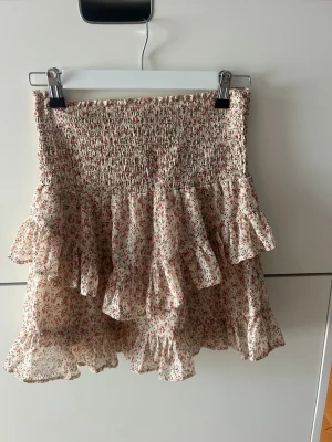 Blommig volangkjol från Neo Noir - Supersöt kort kjol från Neo Noir med blommigt mönster i rött och grönt på beige botten. Kjolen har smock i midjan och flera volanger som ger en flowy känsla. Perfekt för vår och sommar! Storlek S