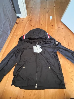  Moncler vindjacka windbraker jacka - Svart vindjacka från Moncler med huva och dragkedja framtill. Jackan har vita och röda ränder på huvan, två fickor med tryckknappar och Moncler-logga på ärmen. Tillverkad i lätt och glansigt material, perfekt för blåsiga dagar.