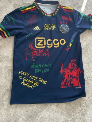 Ajax x Bob Marley limited edition tröja - Unik mörkblå Ajax x Bob Marley limited edition matchtröja från Adidas med färgglada detaljer och tryck. Tröjan har reggaeinspirerade ränder på ärmarna, Ziggo-logga, Ajax-emblem och flera Bob Marley-citat och illustrationer i rött, gult och grönt. Perfekt för fotbollsfans och samlare. Använd 2 gånger ungefär inga defekter ngnstans