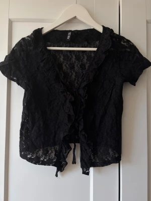 Svart spetsblus från Pieces - Säljer en svart, kortärmad spetsblus från Pieces med volangkant längs öppningen och knyt framtill. Blusen är genombruten med blommigt spetsmönster och har en feminin, croppad passform. Perfekt att styla över ett linne eller topp.