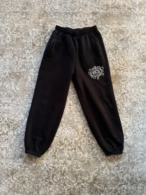 Svarta mjukisbyxor Hugo - Säljer ett par svarta mjukisbyxor med resår i midjan och benslut. Byxorna har två sidofickor och en vit broderad logga på ena benet. Tillverkade i mjukt material som känns skönt mot huden. Perfekta för chill dagar eller när du vill vara bekväm.