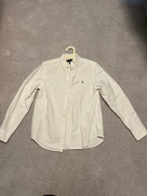 Vit skjorta från Ralph Lauren - Klassisk vit skjorta från Ralph Lauren med broderad logga på bröstet. Skjortan har lång ärm, knappar framtill och en tidlös design som passar till mycket. Tillverkad i mjuk bomull för en bekväm känsla.