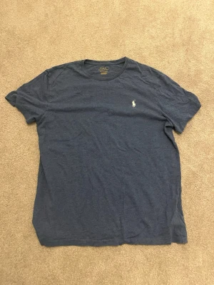 Blå t-shirt från Polo Ralph Lauren - Snygg blå t-shirt från Polo Ralph Lauren med klassisk rund hals och korta ärmar. T-shirten har en liten broderad logga på bröstet och är tillverkad i mjuk bomull. Perfekt för en avslappnad och stilren look.