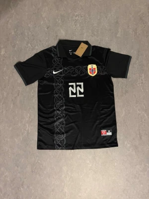 Norge fotbollströja - Snygg svart fotbollströja från Nike med Norges landslagsemblem, nummer 22 och namnet Oscar Bobb på ryggen. Tröjan har korta ärmar, krage och ett unikt mönster längs framsidan. 