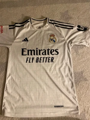 Real Madrid matchtröja Mbappé 9 - Snygg vit Real Madrid matchtröja från Adidas med svarta detaljer och tryck. Framsidan har klubbmärke och sponsorlogga, baksidan har Mbappé och nummer 9. Tillverkad i lätt och ventilerande material med AEROREADY-teknologi. Perfekt för fotbollsträning eller att bära till vardags.