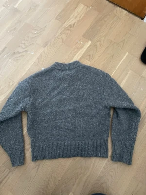Grå stickad tröja - Mysig grå stickad tröja från C med ribbad krage, ärmslut och nederkant. Tröjan har en klassisk rak passform och är perfekt för kyliga dagar. Enkel att matcha med jeans eller kjol för en avslappnad look.