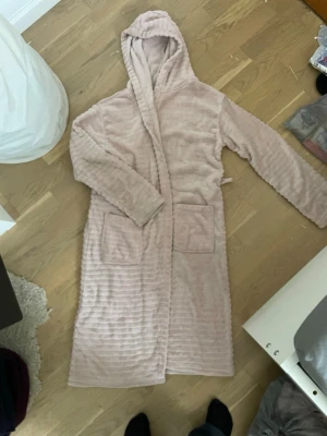 Ljusrosa fluffig morgonrock med huva - Mysig ljusrosa morgonrock i supermjuk fleece med huva och två stora fickor framtill. Perfekt att slänga på sig hemma för extra komfort. Lång modell med skön känsla och enkel knytning i midjan.