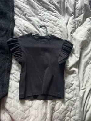 Svart topp med volangärmar - Svart ribbad topp med korta volangärmar och rund halsringning. T-shirten har en slim passform, perfekt för en stilren och trendig look.