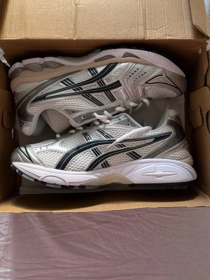 Asics vita och silver sneakers - Snygga sneakers från Asics i vitt och silver med svarta detaljer. Skorna har en sportig mesh-överdel för bra ventilation och klassiska Asics-stripes på sidorna. Tjock, stötdämpande sula och vita skosnören. Perfekta för en trendig och bekväm stil.
