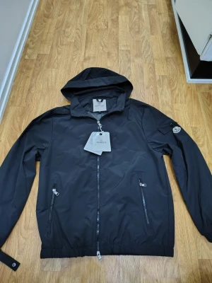 Svart vindjacka från Moncler - Snygg svart vindjacka från Moncler med huva och dragkedja framtill. Jackan har två fickor med dragkedja och Moncler-logga på ärmen. Perfekt för blåsiga dagar och enkel att matcha med olika outfits.