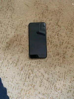 Svart iPhone 7  - Säljer en svart iPhone 7 med klassisk rund hemknapp och stilren design. Baksidan är i matt aluminium och mobilen har en enkel kamera med blixt. Perfekt för dig som gillar Apples smidiga format och snygga look.