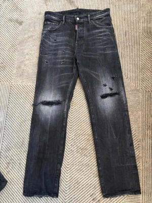 Svarta slitna jeans från Dsquared2 - Säljer ett par svarta jeans från Dsquared2 med snygga slitningar och hål på knäna. Jeansen har raka ben, klassisk femficksmodell och tydliga slitdetaljer framtill. Perfekta för en cool och avslappnad stil.