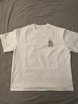  Casablanca t-shirt - Helt ny Vit t-shirt från Casablanca med ett litet tryck på bröstet och ett stort färgglatt tryck på ryggen med texten 'Casa Blanca' och ett motiv av en båge med utsikt. Klassisk passform och rund hals. Perfekt för dig som gillar stilrena men ändå unika plagg.