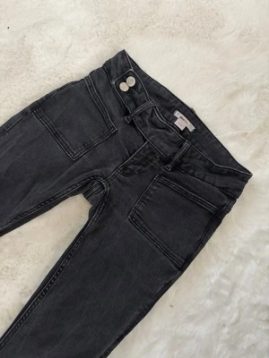 Lågmidjade bootcut jeans - Säljer ett par svarta bootcut jeans med två stora fyrkantiga fickor fram och dubbla knappar i midjan. Jeansen har utsvängda ben och är tillverkade i ett mjukt denimtyg. Perfekta för dig som gillar en retro och avslappnad stil.