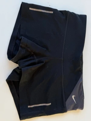 Svarta löparshorts från Nike - Säljer ett par svarta löparshorts från Nike med reflekterande detaljer och Dri-FIT-material som andas. Shortsen har en liten dragkedjeficka bak och en diskret Nike-logga på sidan. Perfekta för löpning eller gymmet.