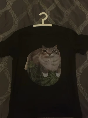 Svart t-shirt med kattmotiv - Cool svart t-shirt med ett stort tryck av en katt på framsidan. Perfekt för dig som gillar djur och vill ha en unik look. T-shirten har klassisk passform och rund hals.