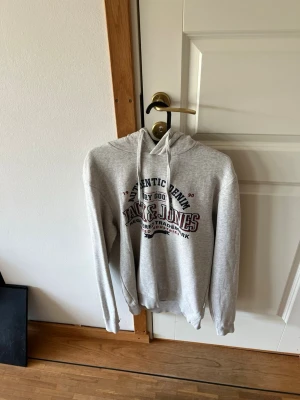 Grå hoodie från Jack & Jones - Snygg grå hoodie från Jack & Jones med tryck på bröstet. Hoodien är i fint skick och sparsamt använd!