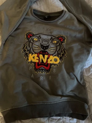 Grå sweatshirt med tigertryck - Cool grå sweatshirt med det ikoniska broderade tigerhuvudet och färgglada detaljer i gult, rött och svart. Tröjan har rund hals, ribbade muddar och är perfekt för dig som vill sticka ut med en trendig streetstyle-look.