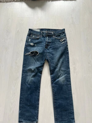 Blå slitna jeans från Abercrombie - Säljer ett par blå jeans från Abercrombie med raka ben och flera slitningar och hål på framsidan. Jeansen har klassisk femficksmodell och är tillverkade i denim med en cool, tvättad look. Perfekta för en avslappnad streetstil.