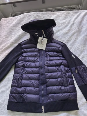 Mörkblå pufferjacka från Moncler - Snygg mörkblå pufferjacka från Moncler med stickade ärmar och vadderad kropp. Jackan har huva, dragkedja framtill, två fickor med knappar och en ärmficka med dragkedja. Moncler-logga på ärmen och coola detaljer i både tyg och glansigt material. Står storlek xl men sitter som M