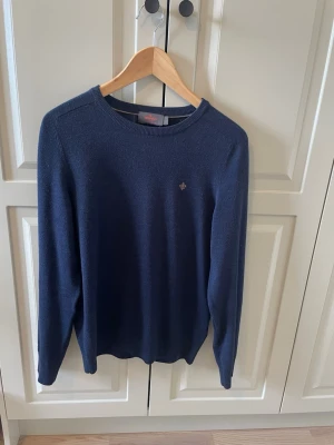 Mörkblå tröja från Morris - Mörkblå rundhalsad tröja från Morris med diskret broderad logga på bröstet. Tröjan har långa ärmar och är tillverkad i ett mjukt, stickat material som passar perfekt till jeans eller chinos. Har en jätteliten öppning som man absolut inte märker av och inte ser när man har på sig tröjan. Därav priset. 