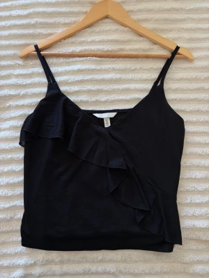 Svart linnetopp med volang från H&M - Svart linnetopp från H&M med smala axelband och snygg volangdetalj framtill. Toppen har även en v-ringning framtill.