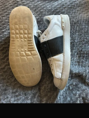 Valentino vita sneakers med svart detalj - Snygga vita sneakers från Valentino med en bred svart läderdetalj över sidan. Klassisk låg modell med rund tå och platt sula. Skorna har diskret Valentino-logga på hälen och är tillverkade i läder för en lyxig känsla.