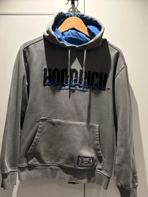 Grå hoodie från Hoodrich med blå detaljer - Snygg grå hoodie från Hoodrich med svart och blå logga på bröstet och blått foder i huvan. Klassisk känguruficka framtill, snörning i huvan och ribbade muddar. Perfekt för en avslappnad streetwear-look.