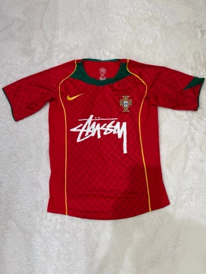 Portugal Nike x Stussy fotbollströja - Röd Portugal fotbollströja från Nike med gröna detaljer vid krage och ärmslut, gula linjer och Stussy-logga på bröstet. Tröjan har broderad Portugal-emblem och är tillverkad i lätt, ventilerande material. Perfekt för dig som gillar streetwear och sport.