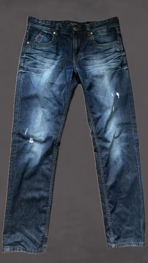 Blå jeans med slitningar från jean biani - Säljer ett par blå jeans med snygga slitningar och tvättade detaljer. Klassisk femficksmodell med raka ben . Jeansen har en casual vibe och passar perfekt till en avslappnad stil.