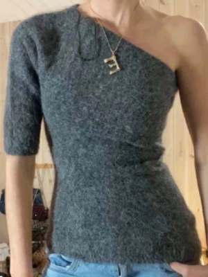 Grå stickad off shoulder tröja - Trendig grå stickad tröja med en axel bar och halvlång ärm på ena sidan. Tillverkad i ett fluffigt, mjukt material som ger en cool och unik look. Perfekt för dig som vill sticka ut med en asymmetrisk stil.