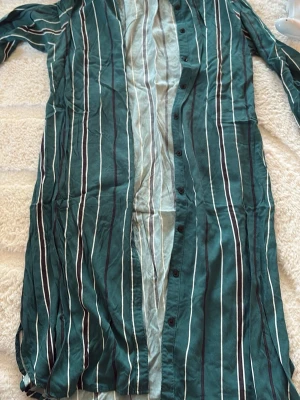 Randig långklänning från Lindex - Snygg långklänning från Lindex i grönt med vita och svarta vertikala ränder. Klänningen har knappar längs hela framsidan och är tillverkad i ett lätt och luftigt material, perfekt för varmare dagar. Klassisk krage och lång ärm för en avslappnad men stilren look.